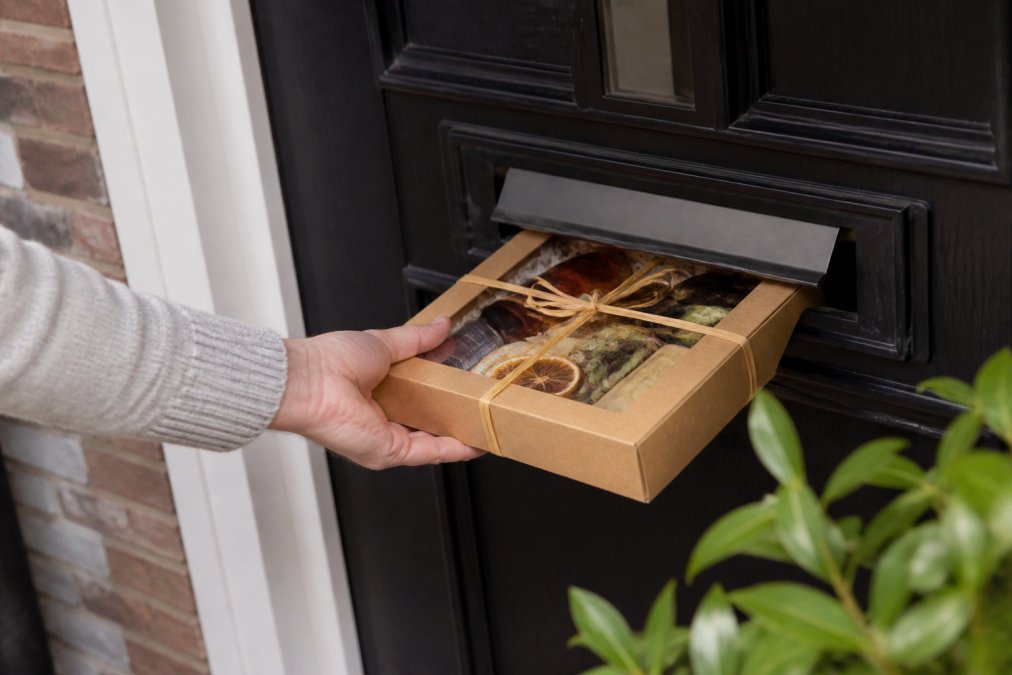 A hand posting a beautifully wrapped Letterbox Cocktails gift box through a standard front door letterbox, with a glimpse of the premium bottles and garnishes visible inside the box
