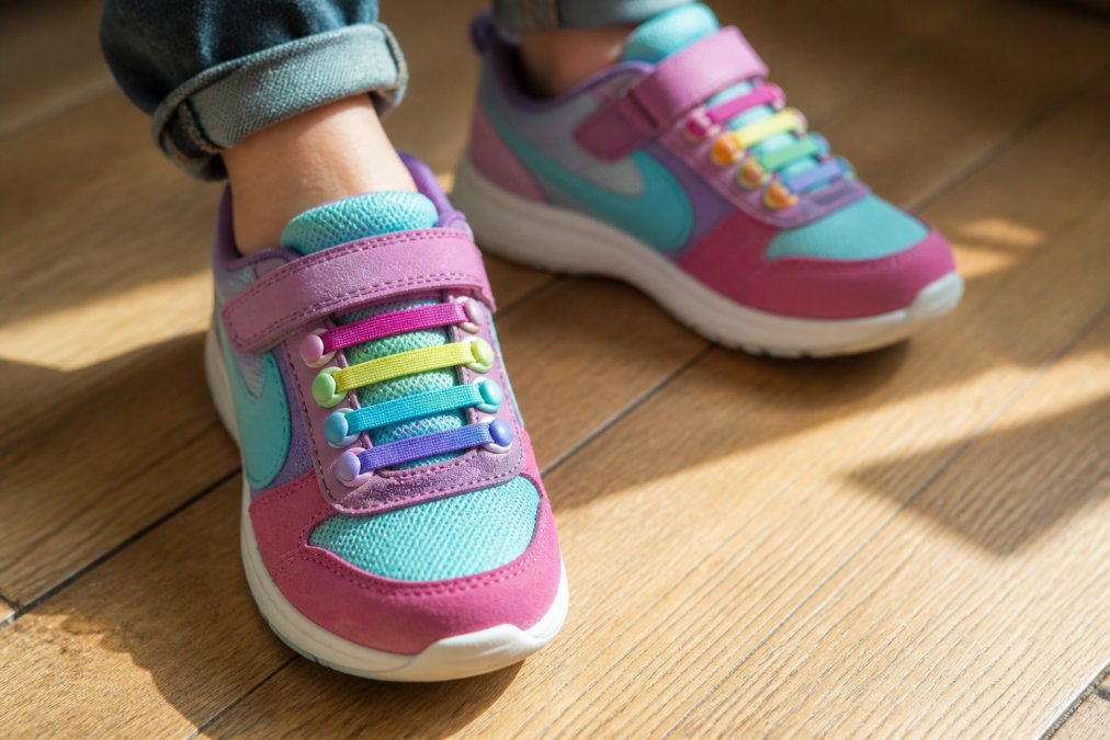 Close-up of a child's colorful sneakers with vibrant elastic no-tie laces threaded through the eyelets, laid flat on a bright wooden floor in warm natural light