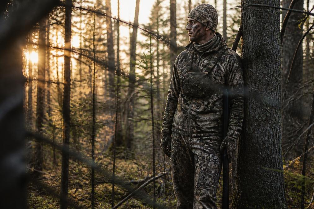 Hunter in full camouflage gear standing motionless in dense wooded terrain during early morning light, wearing layered technical hunting apparel with visible moisture-wicking base layer and insulated mid-layer