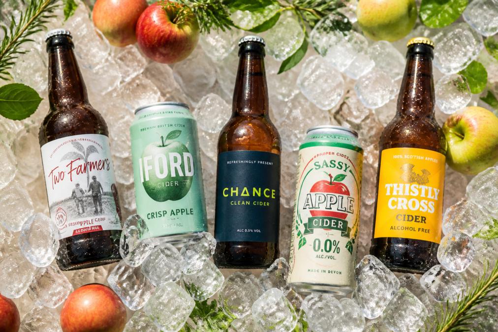 Overhead shot of five different alcohol-free cider bottles and cans chilled on ice with fresh apples and summer herbs scattered around, bright natural sunlight