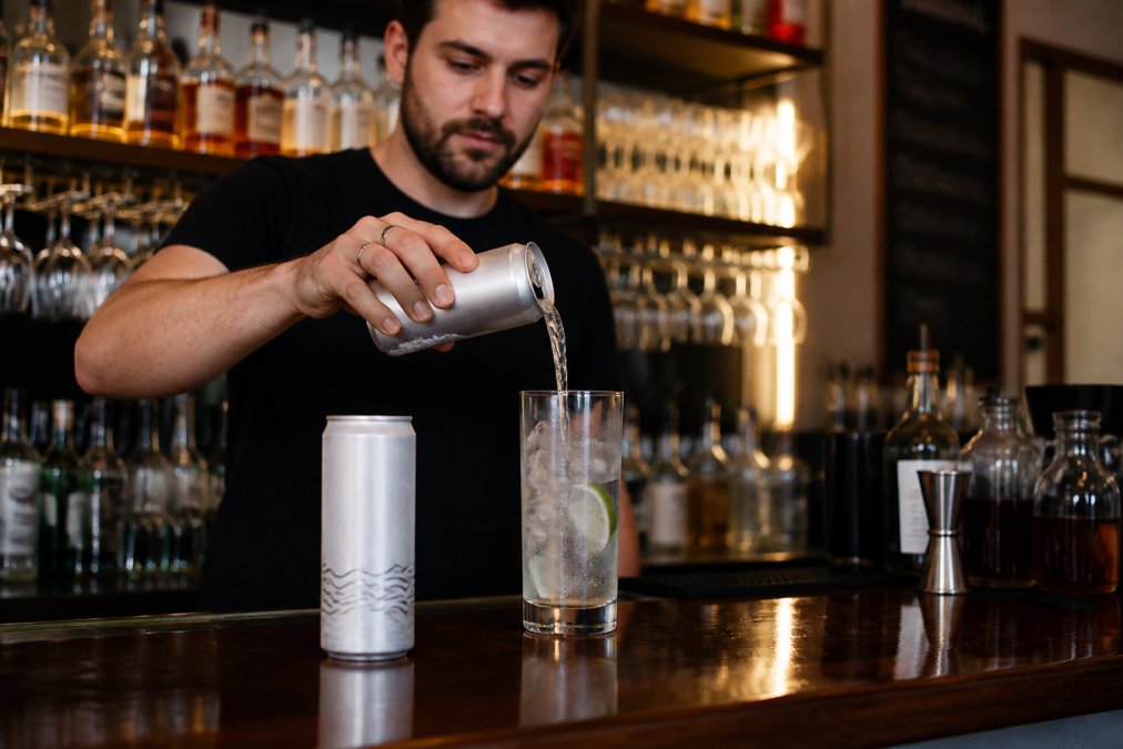 A bartender in a modern, stylish pub pouring a pre-mixed Savyll cocktail from a can into a chilled highball glass with ice and a lime wedge, the product can visible in the foreground on the bar