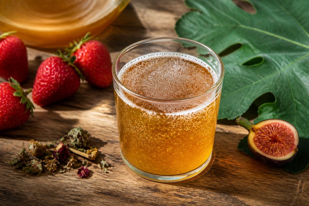 Close-up of a glass of raw kombucha with visible bubbles and fermentation, surrounded by fresh strawberries, fig leaves, and botanicals on a wooden table with natural sunlight