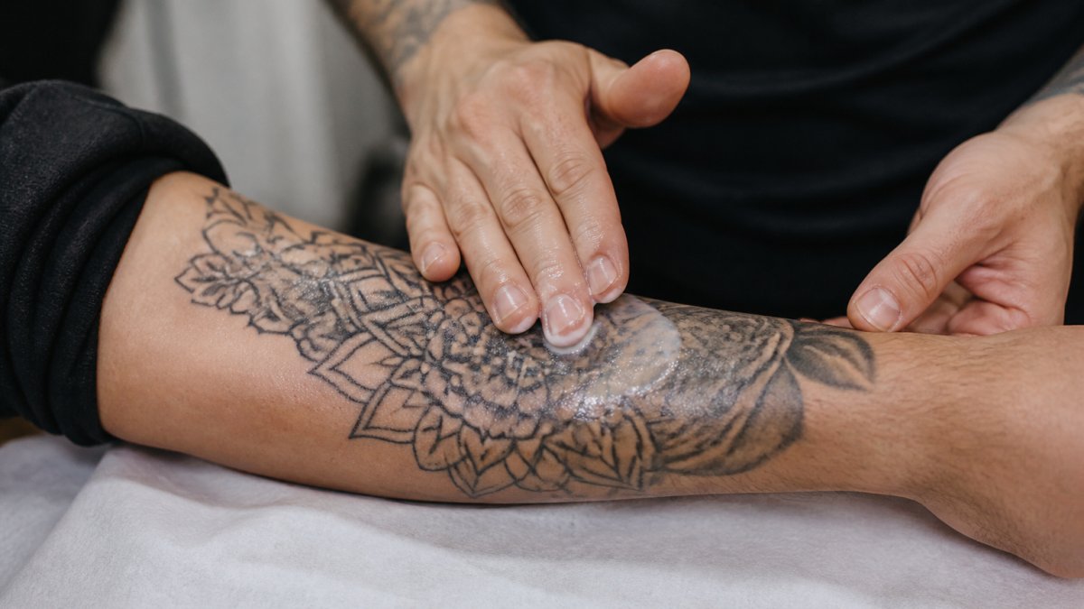 Close-up of hands applying a thin layer of balm to a fresh tattoo on the forearm, showing proper massage technique with circular motions on clean, dry skin