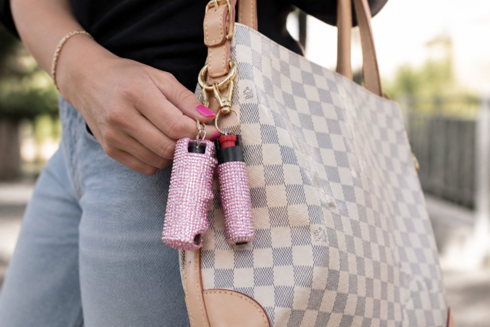 Woman's hand holding a stylish keychain set with rhinestone-decorated stun gun and pepper spray attached to designer purse strap in natural daylight