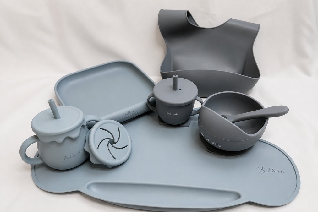 Flatlay of colourful silicone feeding sets arranged on a white surface, showing bowls, plates, bibs, and utensils from different sets with natural daylight