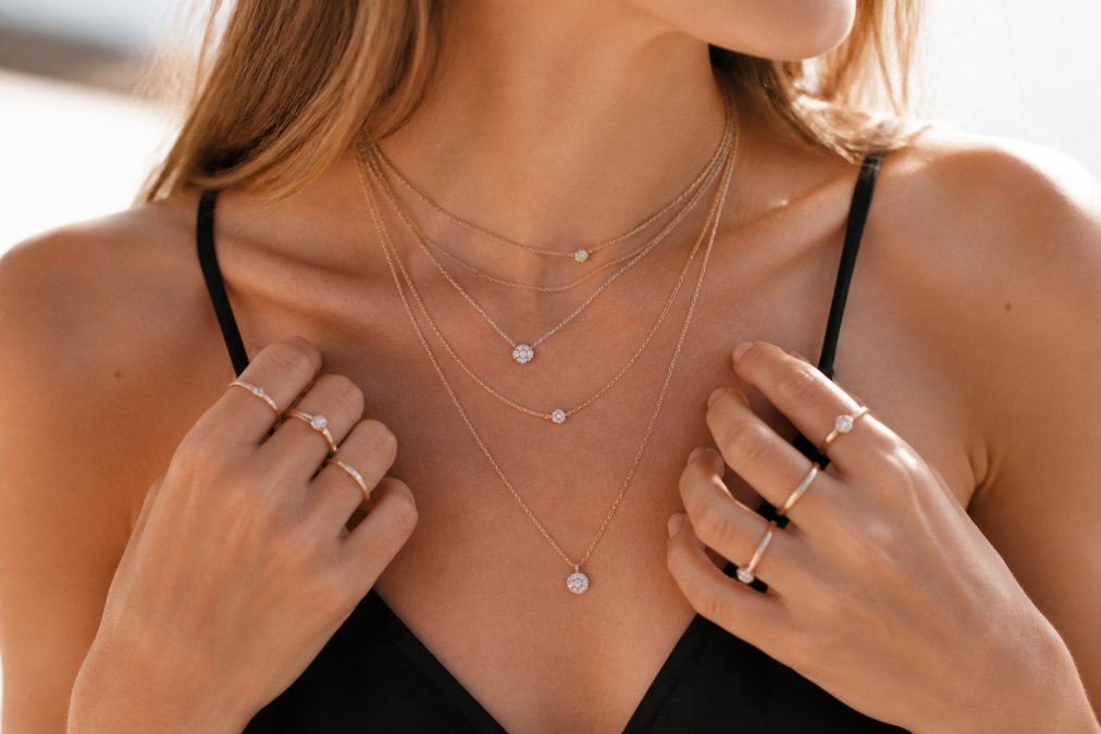 Close-up of hands displaying multiple thin stacked rings and delicate layered necklaces in various metals and finishes, demonstrating versatile everyday jewelry combinations