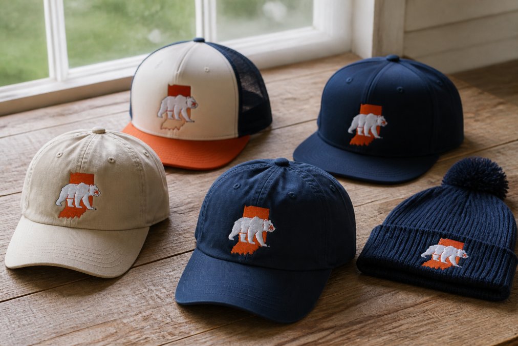 Collection of Indiana Bears hats arranged on a wooden surface showing variety, including classic dad cap in khaki, mesh-back trucker cap, flat bill snapback with embroidered logo, and knit beanie, photographed in natural window lighting