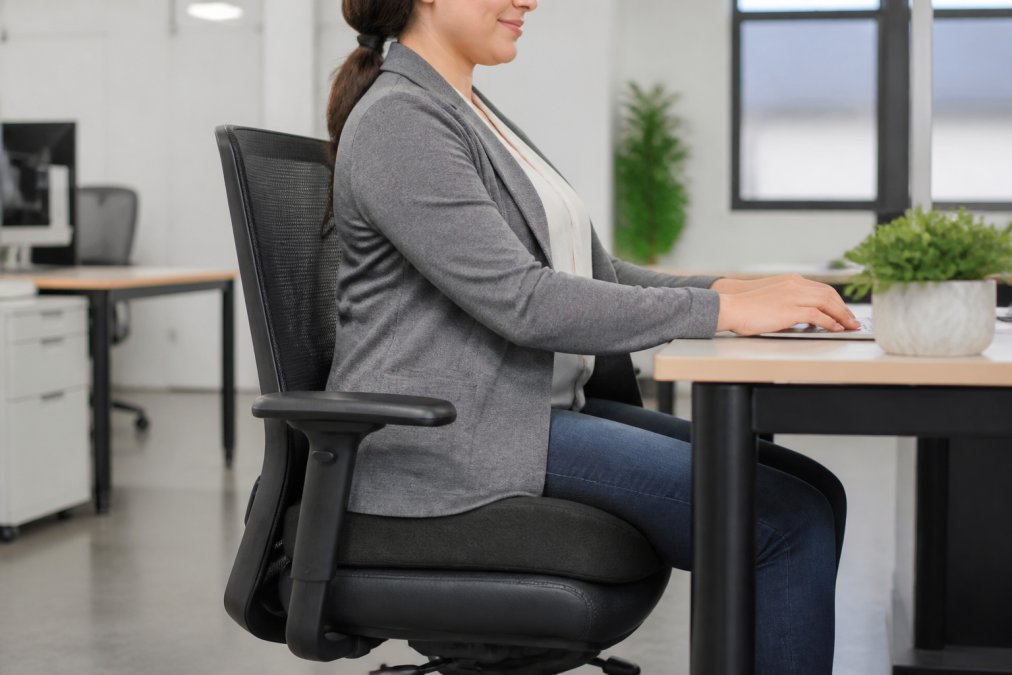 An office worker seated at a desk with a contoured seat cushion visible on their chair, showing a relaxed posture in a bright modern workspace