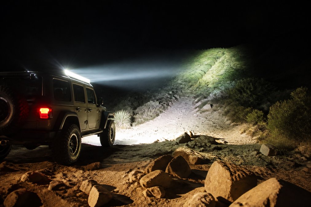 Off-road vehicle at night with LED light bar mounted on roof, showing distinct spot beam reaching far into darkness illuminating distant terrain and flood beam lighting nearby trail, rocky obstacles visible in foreground