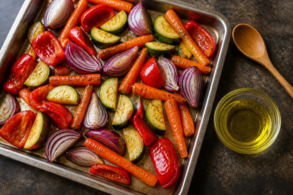 An overhead shot of a baking sheet filled with vibrant carrots, red bell peppers, red onion wedges, and zucchini, all glistening with a light coating of oil, with a small glass bowl of golden avocado oil and a wooden spoon resting beside the tray on a dark countertop