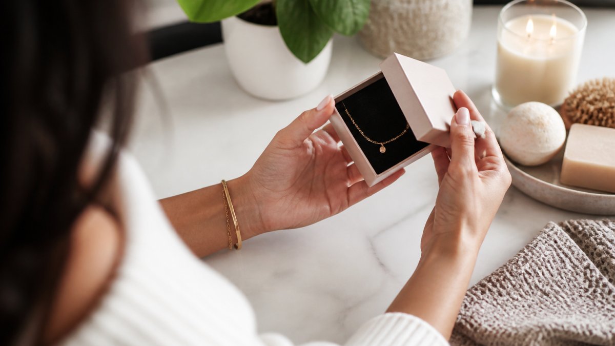 A woman's hands holding a small jewelry gift box with a delicate gold bracelet inside, soft natural light on a marble countertop, a lit candle and small plant nearby