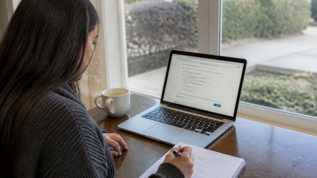 A person sitting at a desk with a laptop showing a practice test, a notepad with handwritten notes, and a cup of coffee nearby
