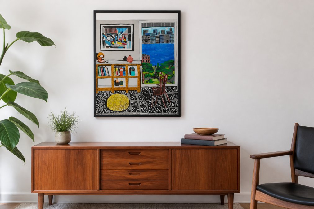 A contemporary art print in a thin black frame mounted on a white wall above a mid-century modern sideboard, with a small plant and books styled nearby in a minimal living room