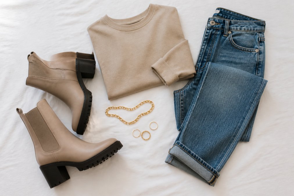 A neatly arranged flat lay of versatile wardrobe staples including a pair of chunky ankle boots, a neutral beige top, straight-leg jeans, and minimal gold accessories on a clean white background