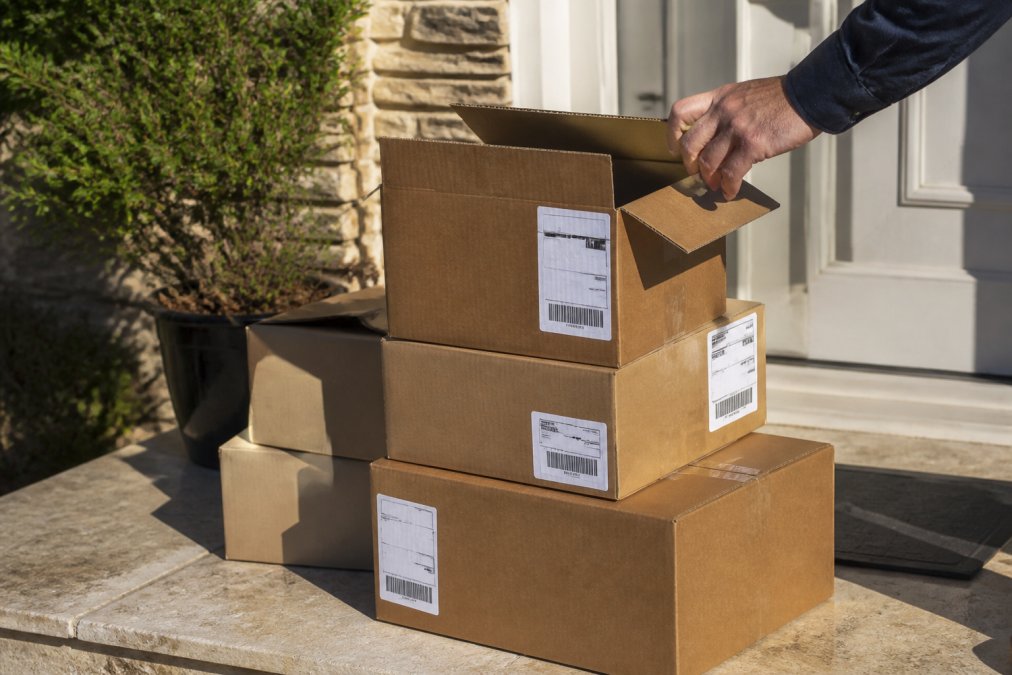 A stack of cardboard delivery boxes with different shipping labels sitting on a sunlit doorstep, one box being opened by a hand