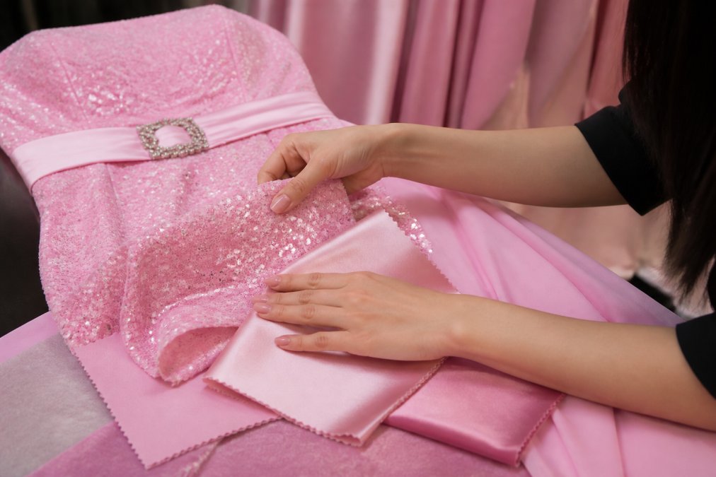 Woman's hands touching premium sequined and satin fabric samples, fingers tracing the light-catching micro-sequins and smooth satin weave, showing texture detail and shimmer under warm studio lighting