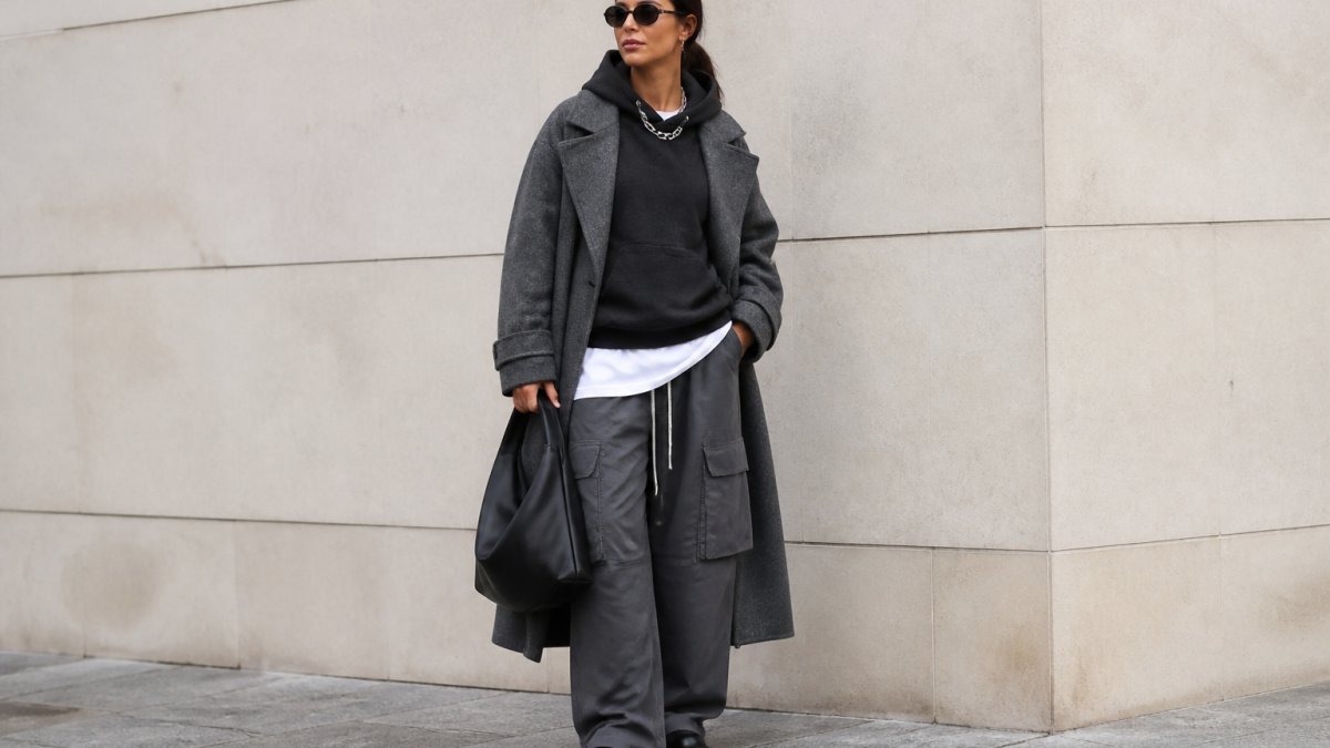 A confident woman in her 30s wearing a bold, layered streetstyle outfit standing against a clean urban wall, natural daylight, full-body shot showing the outfit's texture and silhouette