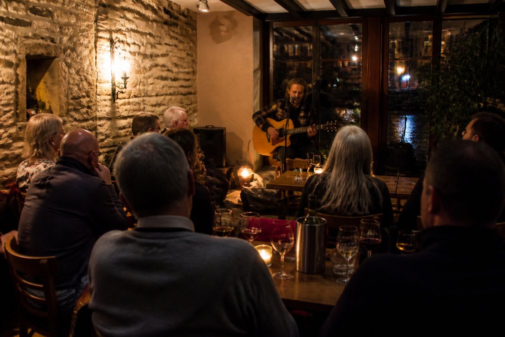 Intimate riverside bistro with warm lighting, performer with acoustic guitar on small stage, audience seated at tables with wine glasses, historic stone building interior