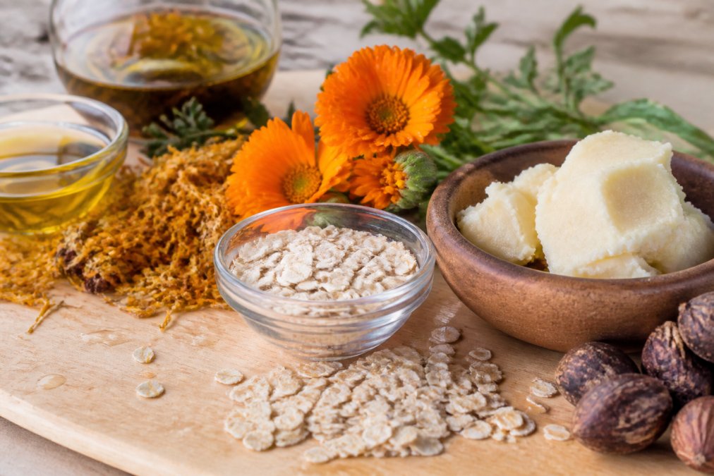 Close-up of natural organic ingredients including colloidal oatmeal, shea butter, calendula petals, and botanical extracts arranged on a light wooden surface with water droplets glistening in soft natural light, showing texture and purity
