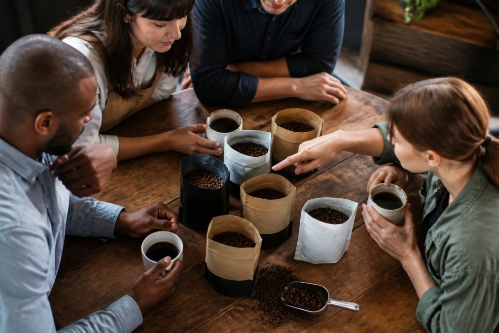Overhead shot of various whole bean coffee bags with visible origin labels and roast profiles, arranged on a wooden surface with a coffee scoop and freshly roasted beans scattered nearby