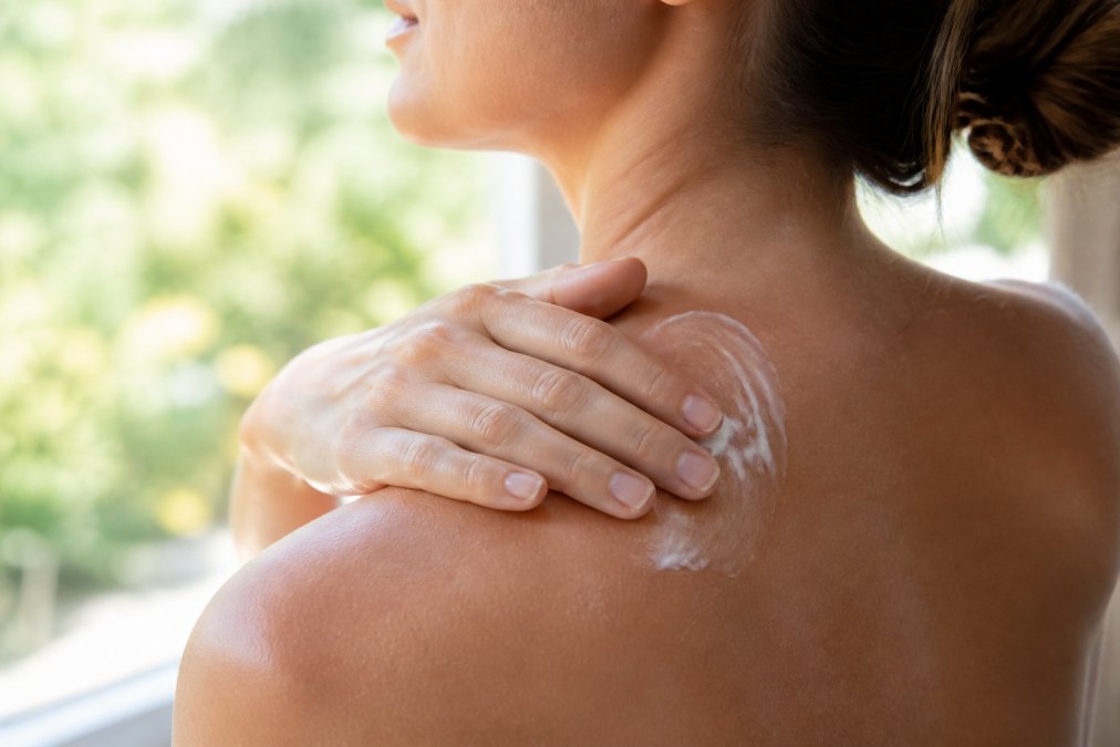 Close-up of hands applying a rich, non-greasy cream to shoulder and neck area, showing the texture and absorption of a botanical skincare product in natural light