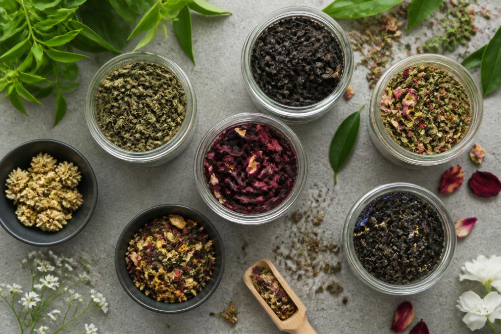 Overhead flat lay of loose leaf tea varieties in glass jars and bowls, showing different colors and textures of green, black, and herbal teas with fresh leaves and flowers scattered around