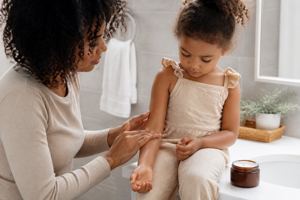 A parent gently applying organic eczema cream to a young child's arm in a softly lit bathroom, with a jar of botanical skincare product on the counter nearby
