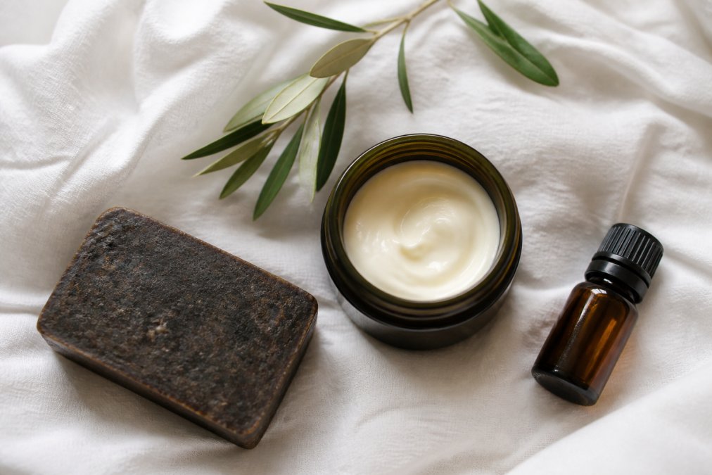A flat lay of organic skincare products including a bar of dark African black soap, a jar of healing cream, and a small bottle of essential oil on a clean white linen background with soft natural light