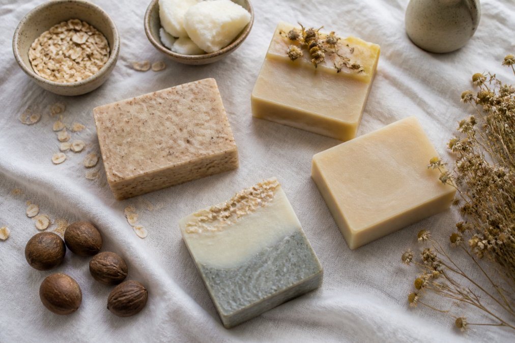 Flat lay of several artisan cold-process soap bars with natural ingredients like oats, shea butter, and chamomile flowers arranged on a white linen cloth in soft natural light