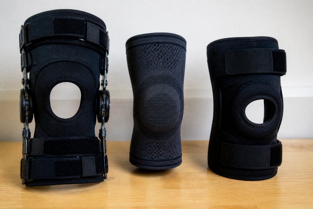A clean, organized display of several different types of knee braces laid out on a neutral background: a rigid hinged brace with aluminum stays, a knit compression sleeve, and a wrap-around brace with straps