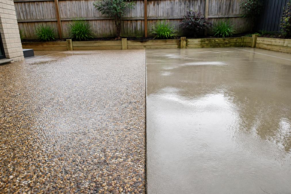 A close-up photograph of a smooth pebble paving surface on a residential patio, with a garden bed and a low timber fence in the background, taken on a bright day after rain