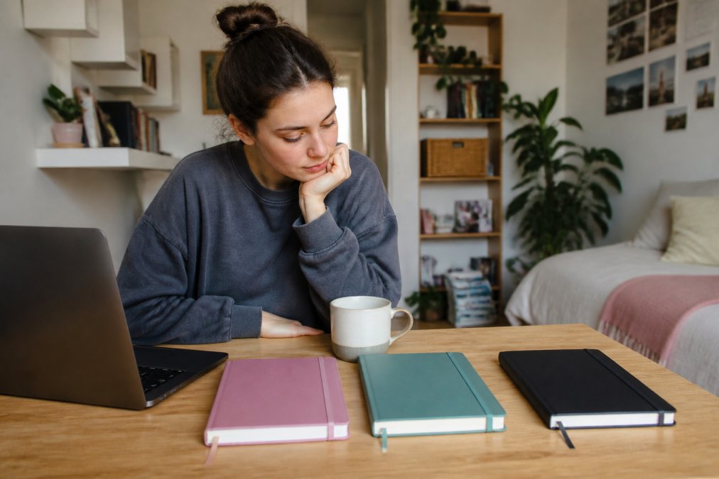 A college student sitting at a wooden desk with several journals, a laptop, and a cup of coffee, reviewing options in a bright dorm room