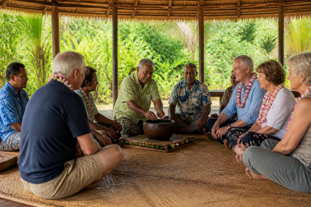 A small group of mature travellers seated on woven mats inside a traditional Fijian bure, sharing a kava ceremony with village elders, warm natural light streaming through the open sides, lush tropical greenery visible beyond