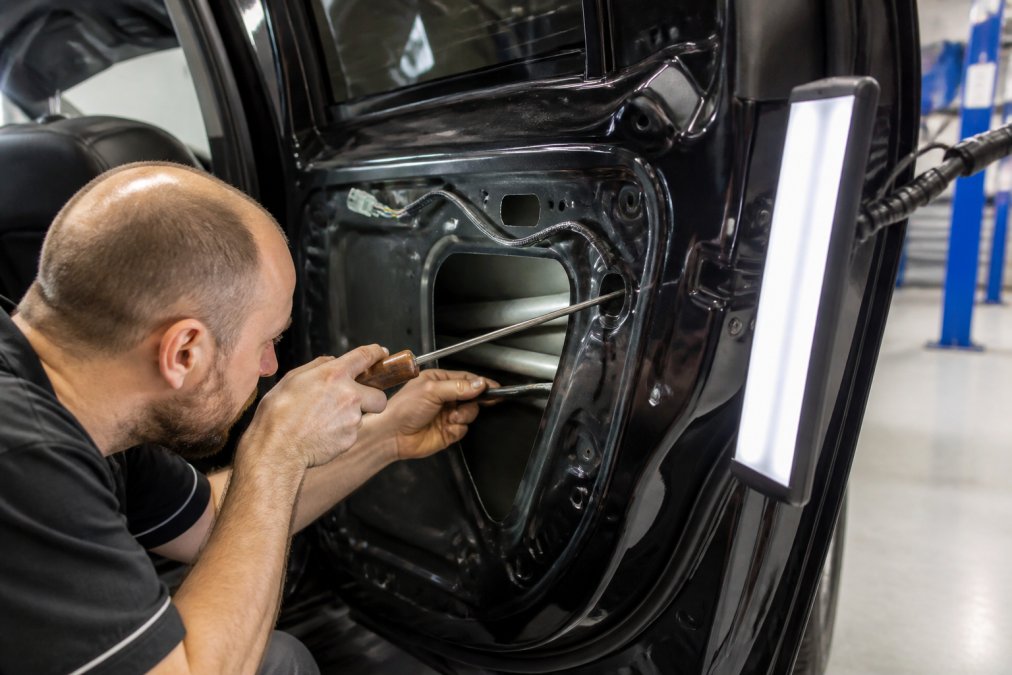A professional paintless dent repair technician using specialized rods and hooks to gently massage a hail dent from the inside of a car panel, with the vehicle's factory finish clearly visible and a clean, well-lit shop environment in the background
