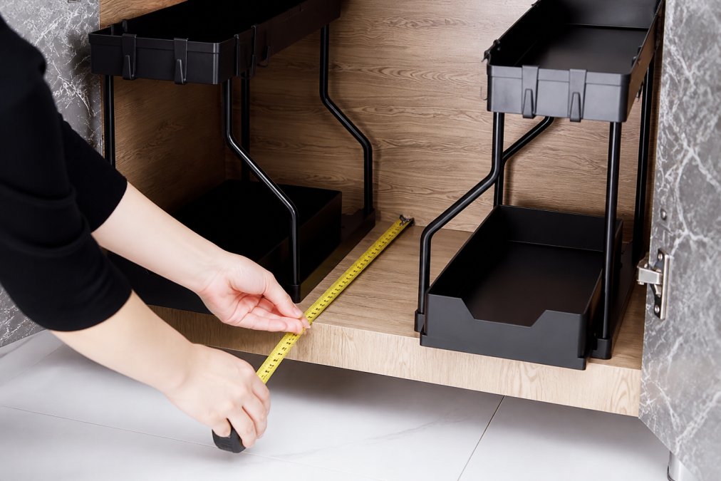 Close-up of hands holding a tape measure against a deep wooden pantry shelf, measuring the depth from front edge to back wall with clear measurement markings visible in natural kitchen lighting