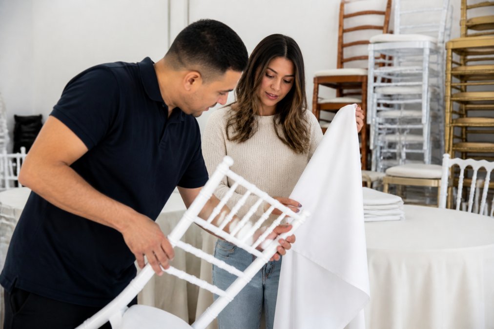 Close-up of hands carefully inspecting white resin chair frames and polyester linens for cleanliness and structural integrity, with a rental company representative explaining quality standards in a bright showroom setting