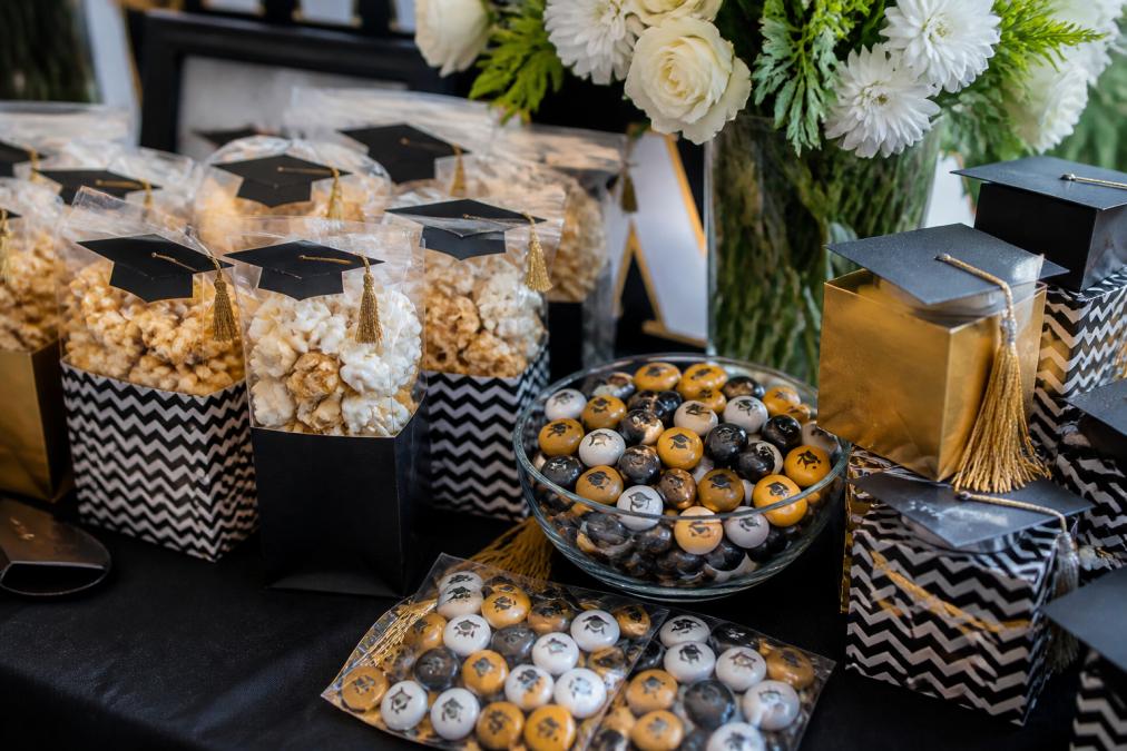 Close-up of personalized graduation party favors including custom popcorn bags with school colors, personalized M&M candies in branded packaging, and decorative keepsake boxes arranged on a table with graduation decorations and flowers