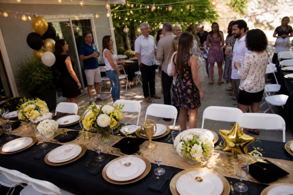 Overhead view of a graduation party setup with coordinated tableware, colorful balloons, centerpieces, and guests mingling around a decorated outdoor space with string lights and banners