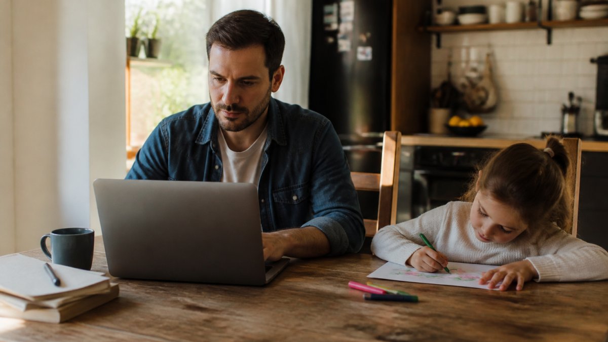 A parent working on a laptop at a kitchen table with a child's drawing and coffee cup nearby, warm morning light