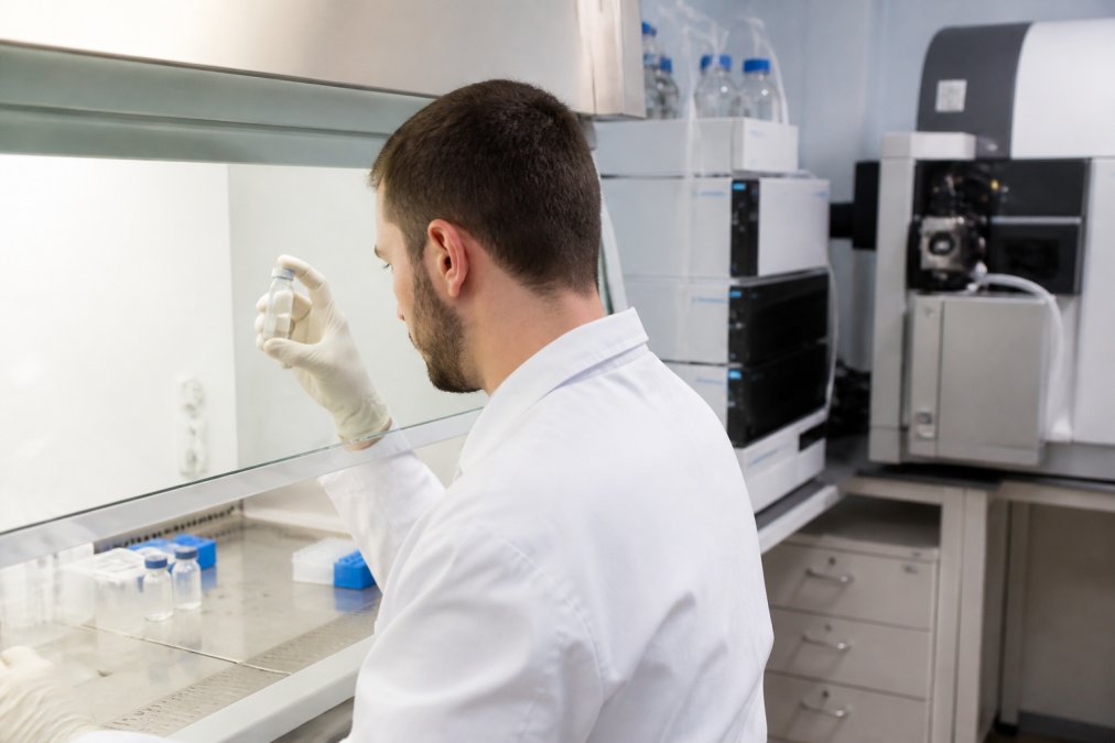 Laboratory technician in white coat examining a clear vial of peptide sample under a fume hood with HPLC and mass spectrometry equipment visible on the bench in the background