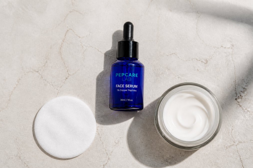 Overhead flat-lay of skincare products arranged on a light background, showing a serum bottle, cream jar, and cotton pad in a simple routine setup