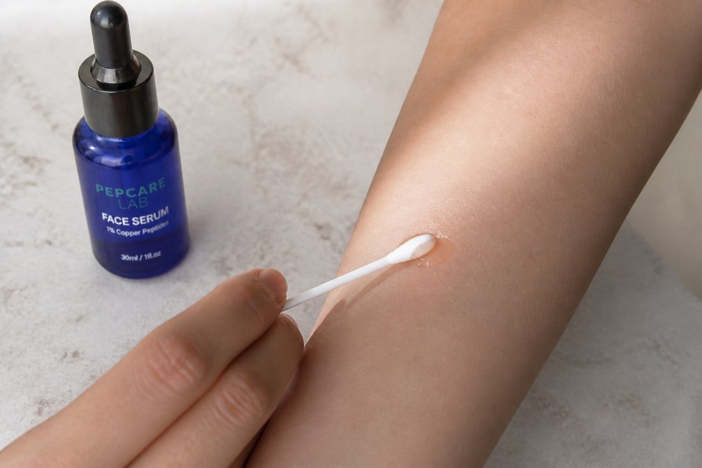 Close-up of a person's forearm showing a small amount of serum being applied to a test area with clean, natural lighting and a cotton applicator