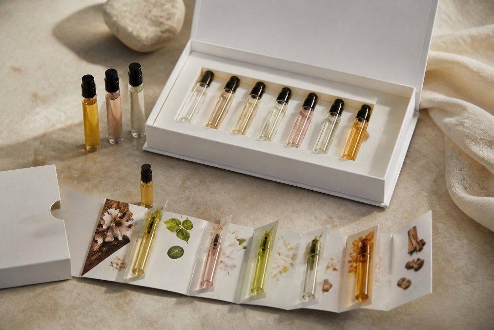 Artfully arranged Ruth+Bo perfume discovery set with opened luxury box, multiple mini atomizer bottles displaying various fragrance colors, sample cards with scent descriptions, arranged on a neutral surface with soft natural lighting