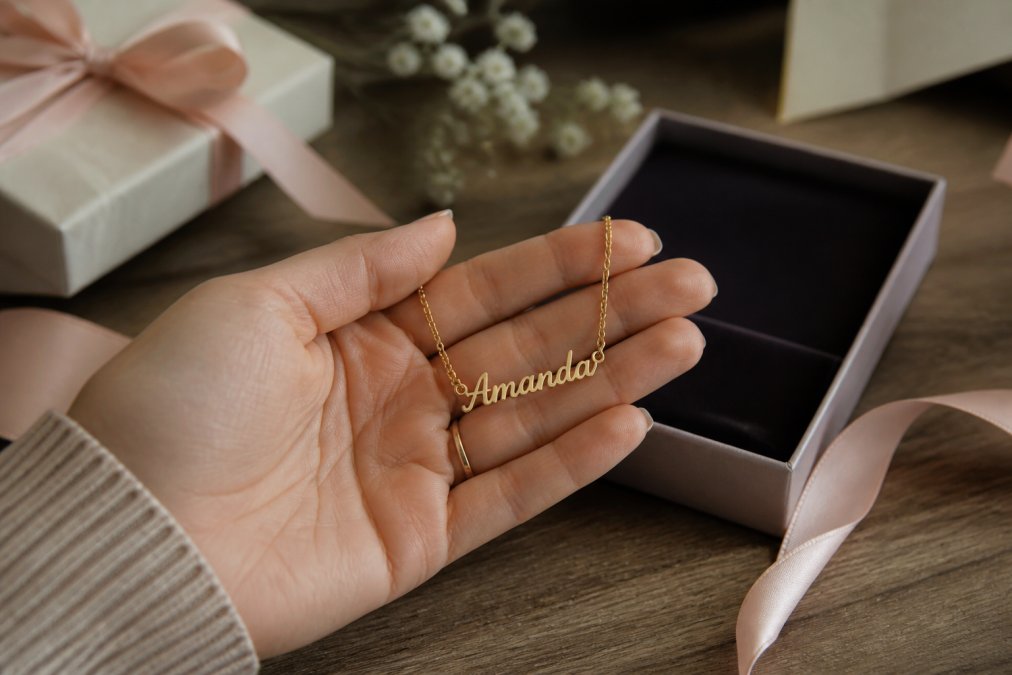 A close-up of a woman's hand holding a delicate personalized name necklace, with a soft-focus background of an open gift box and elegant wrapping paper on a wooden table