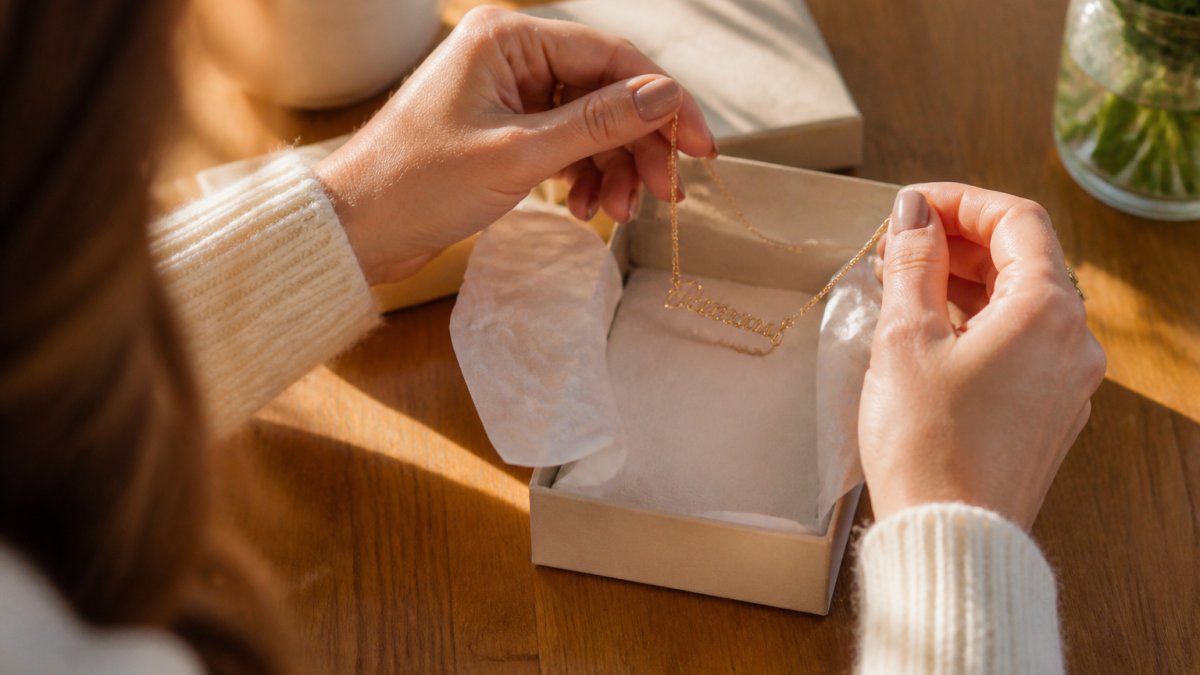 Close-up of a woman's hands lifting a delicate gold cursive name necklace out of a soft-touch gift box lined with tissue paper, warm window light across a wooden table, sentimental gifting moment
