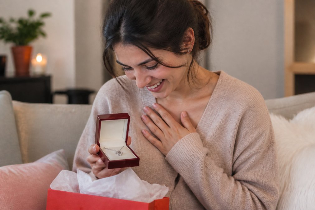 Woman opening a beautifully wrapped gift box containing a personalized love knot necklace, smiling with joy and emotion as she holds the jewelry piece close to her heart