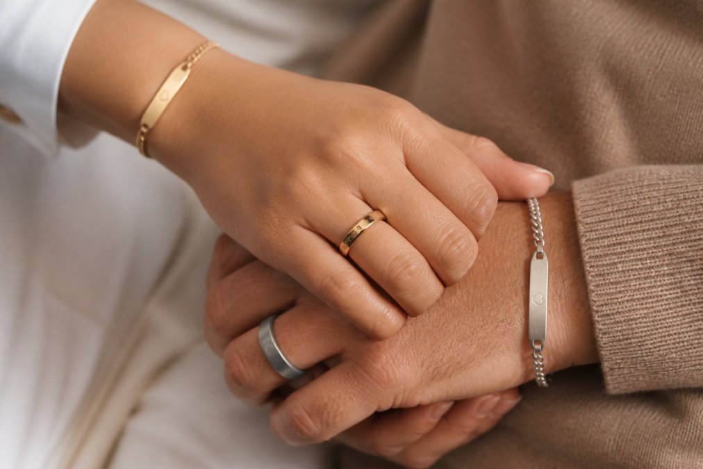Couple's hands together displaying matching personalized rings and bracelets with engravings, warm lighting emphasizing the sentimental moment