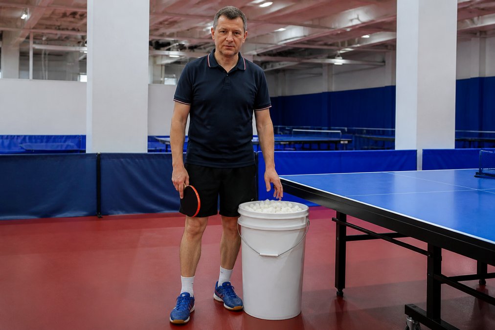 A coach standing upright beside a plastic bucket full of white ping pong balls next to a table tennis table in a bright training hall