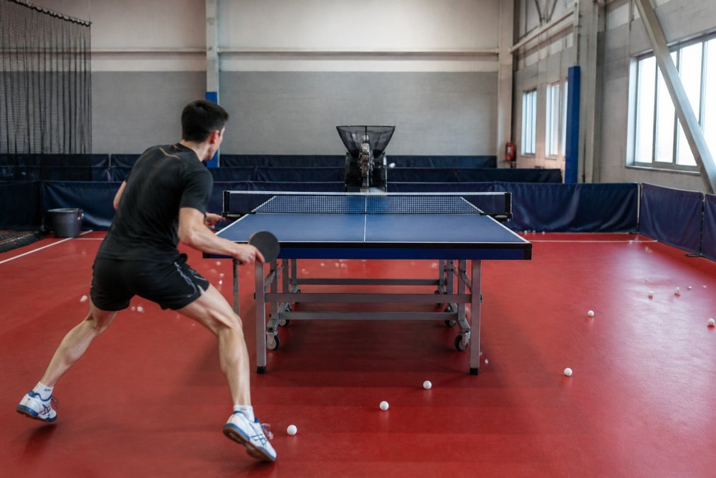 A solo player in a well-lit indoor sports facility returning balls from a table tennis robot mounted at the far end of the table, footwork visible as they move laterally across the court