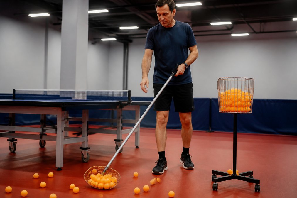 A coach standing upright using a telescoping ball collector to scoop scattered orange ping pong balls from a gym floor beside a table tennis table, bright overhead lighting in an indoor sports facility